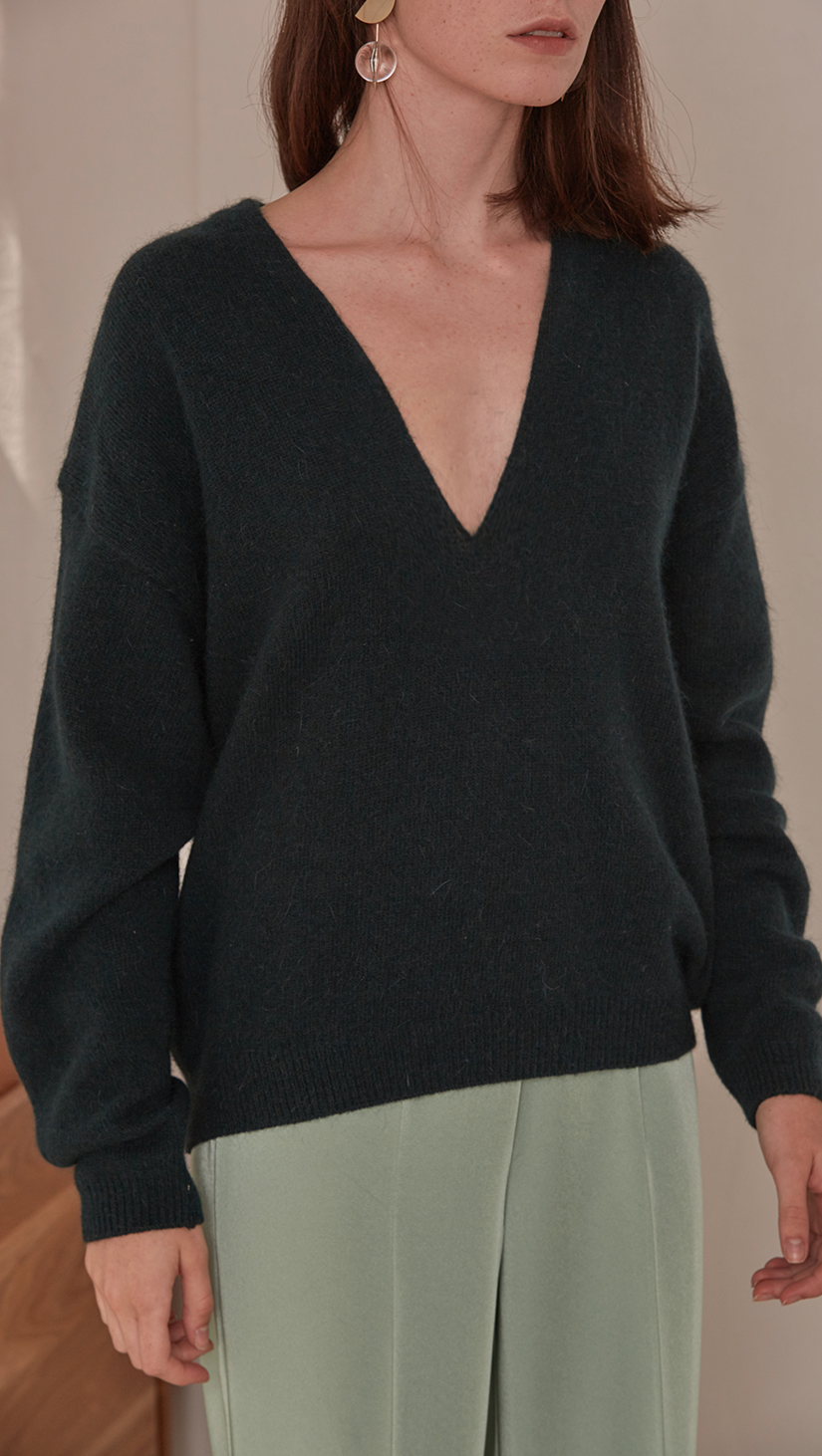Agostina Sweater in Dark Green. Long sleeved pullover with deep v-neck in dark green lambs wool blend. Cowl neck ribbed sweater with pointed dropped shoulder seams and extra long sleeves. Designed to be loose fit.