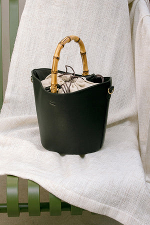 Bonet bag in Black. Wooden bamboo carrying top handles. Cylindrical design. Brown drawstring fabric insert with white tassel.