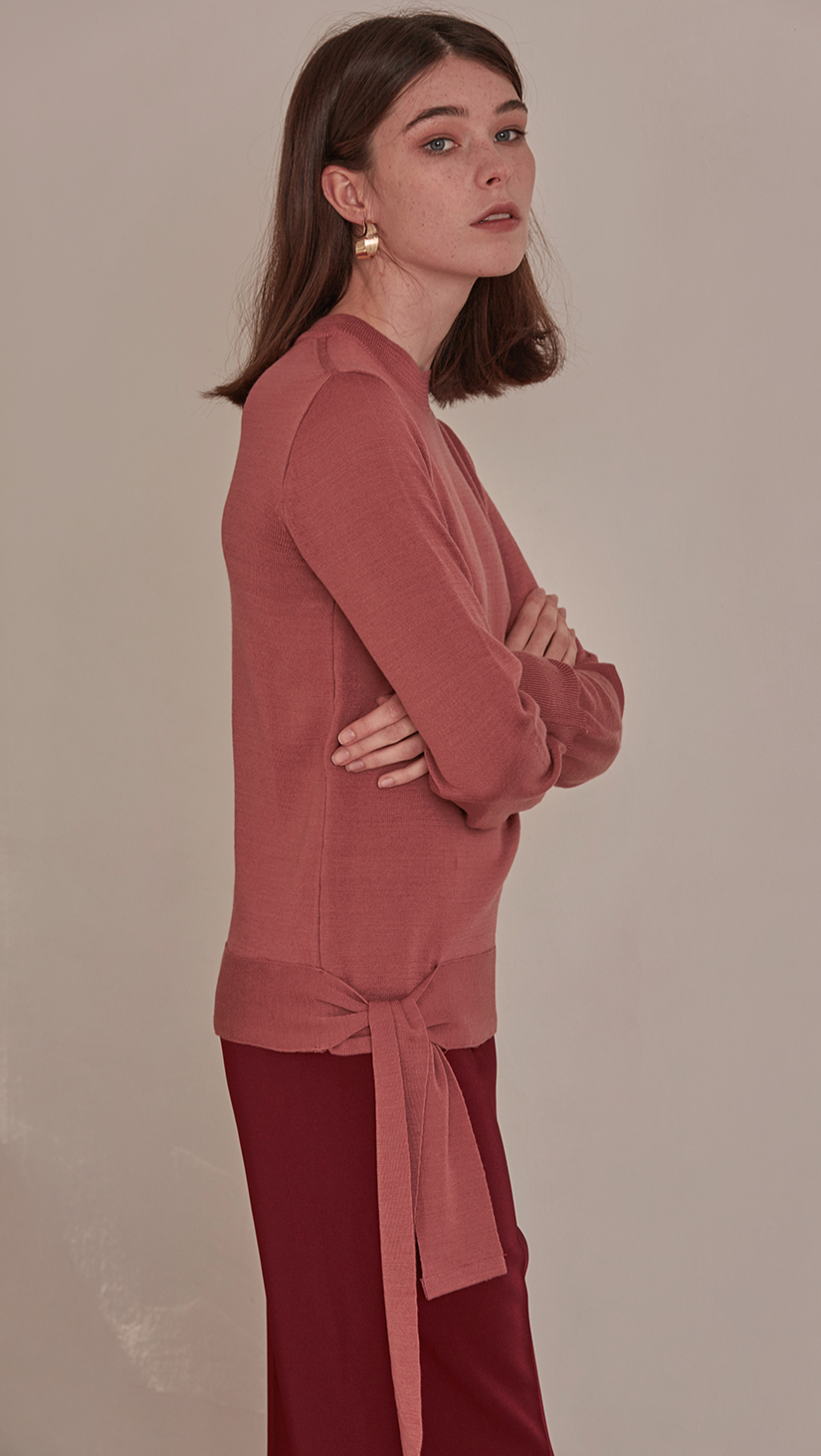 Féte Sweater in Pink. Crewneck long sleeved lightweight knit. Self wrap tie on the side of hem. Slit at cuff. Pull on. Slim fitting body with a long tie sash. 