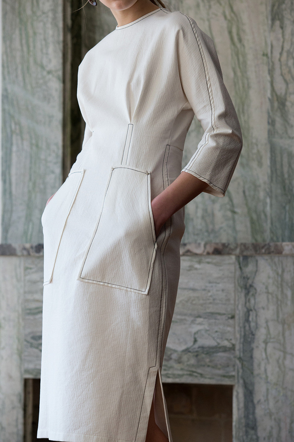 The Kaveh Dress in White, featuring three-quarter sleeves with stitching details, ruched at waist, two slant pockets. Concealed zip fastening at the back. Fitted at waist. Side slits.