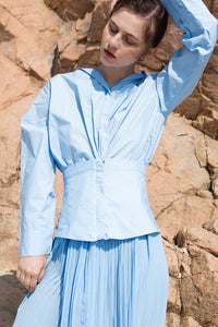 The Leslie Dress in Blue featuring stylized snap button-down shirt dress. Wide v-neckline, spread collar, full-button placket with twisted detailing. Detachable A-line shaped wide belt with concealed zip closure at waist. Dropped shoulder. Long bell sleeves with wide button cuff. Long silhouette. Oversized fit.