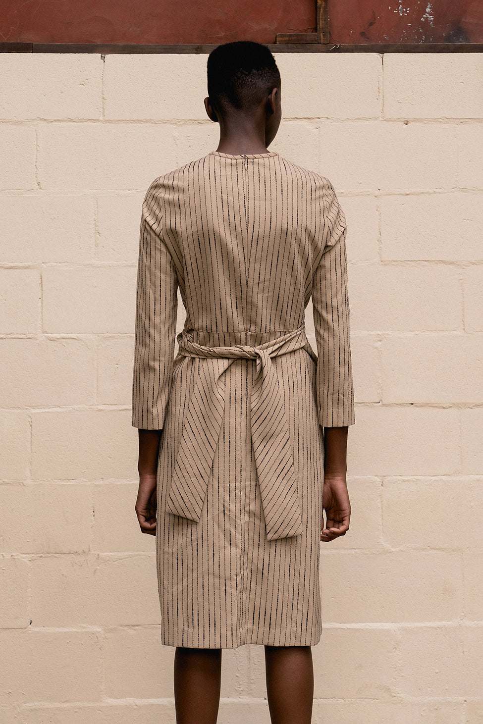 The Lorraine dress featuring round neckline, three-quarter sleeves, gathered pleats at front with self-tie. Concealed zip closure at back. Unlined. Mid-length. 
