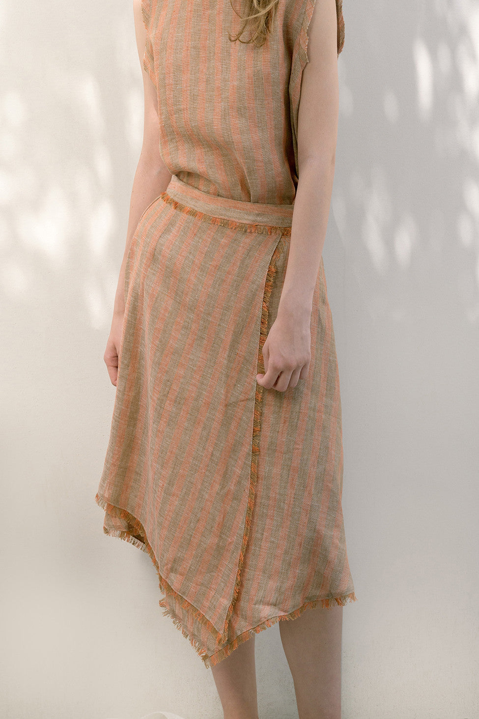 The Majira Skirt in Orange featuring asymmetric wrap design in fluted raw edge with tassel detailing. Concealed side zip closure. High waist in below-the-knee length. Partial lined.