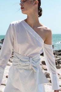 The Roslyn Top in white featuring one shoulder with long sleeves, pearl hardware button at cuffs, elastic band at back. Pull on.
