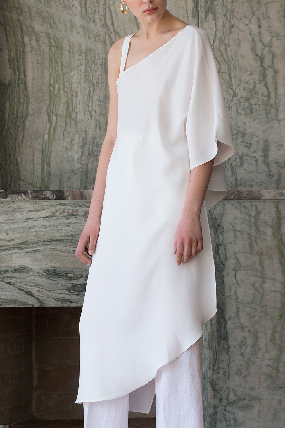 The Rowe Tunic in White. Cut out at one shoulder with D-ring adjustment. Asymmetric hem. Semi sheer. Pull on. 