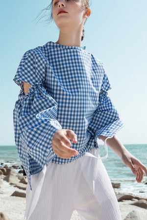 The Tabitha Top in blue gingham stripes featuring boat neckline, open-back with drawstring tape back tie. Gathered long wide sleeves. 